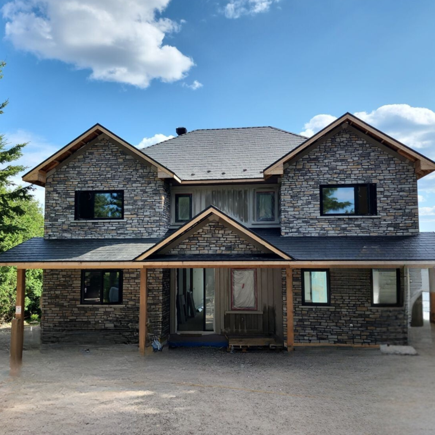 Maison en construction avec murs en pierre, fenêtres à battants, et toiture en pente sous un ciel bleu avec nuages.