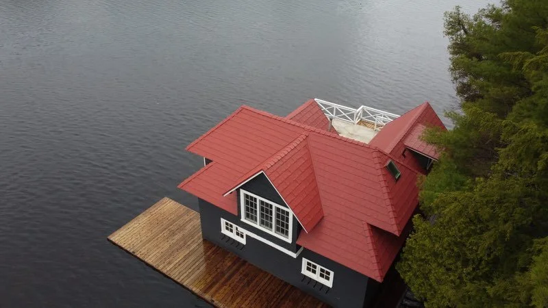 Une maison en bois gris avec un toit rouge située au bord d'un lac bordé d'arbres.
