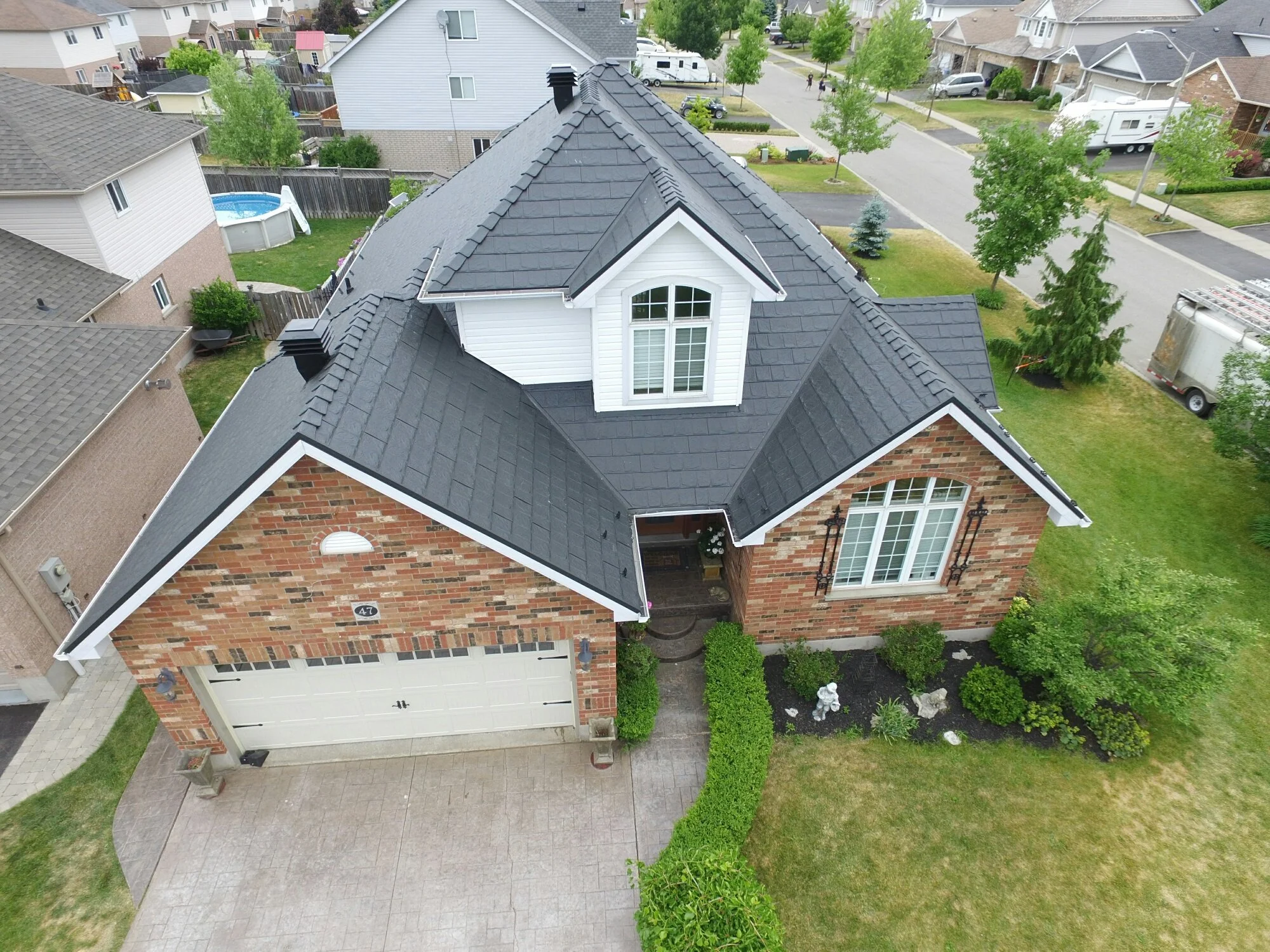 Vue aérienne d'une maison résidentielle en briques avec un toit en ardoise noire, un garage, un petit jardin avec des buissons et un chemin menant à l'entrée principale, dans un quartier résidentiel calme avec des arbres et d'autres maisons voisines.