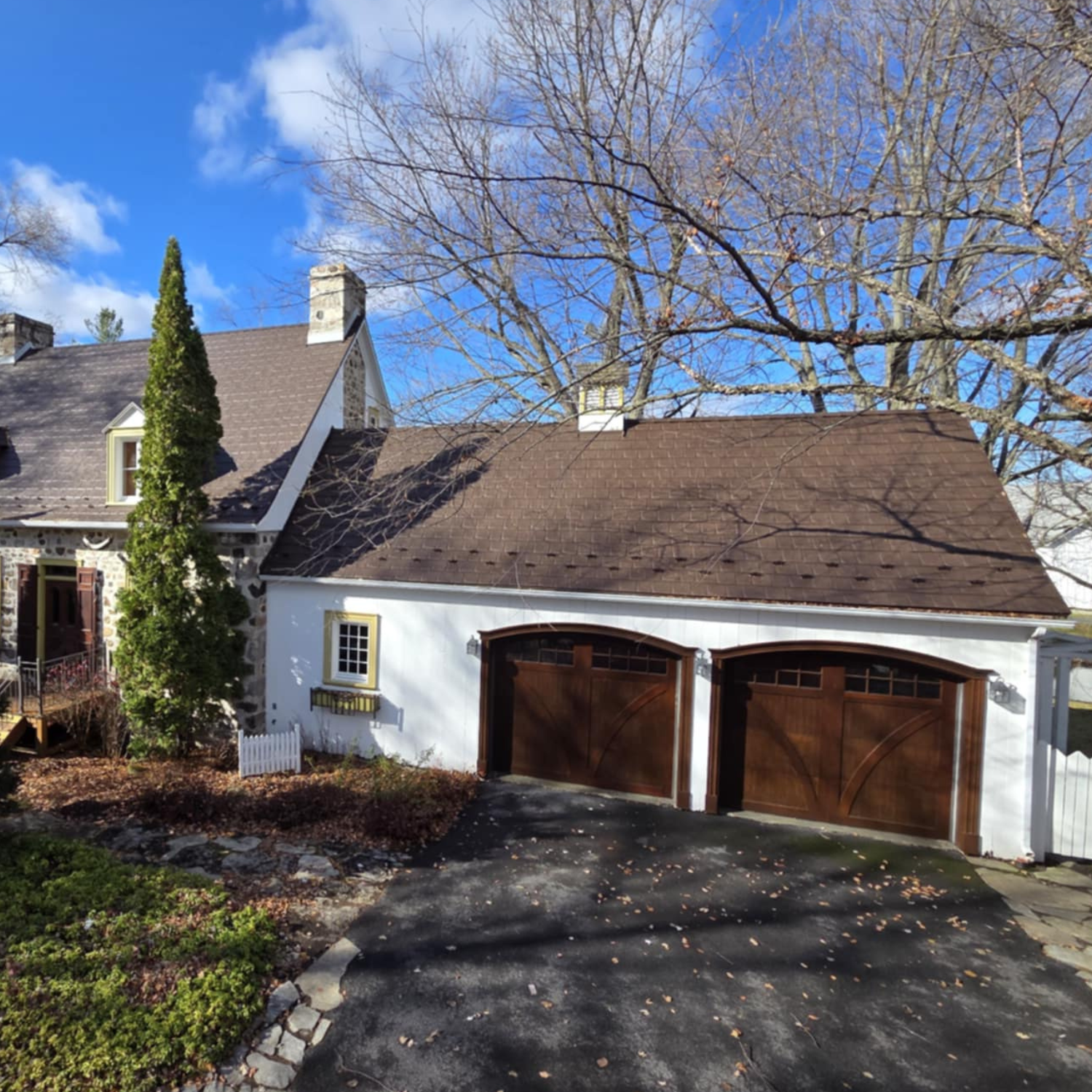Une maison avec un garage à deux portes en bois, un arbre sans feuilles en arrière-plan, et un ciel bleu avec quelques nuages.