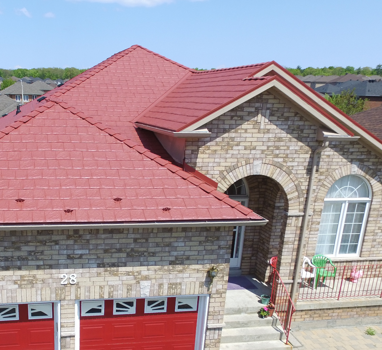 Maison en briques avec un toit rouge et une fenêtre avec des rideaux, entrée principale avec une petite terrasse, place pour deux chaises vertes et une chaise blanche, numéros 28 sur la porte de garage.