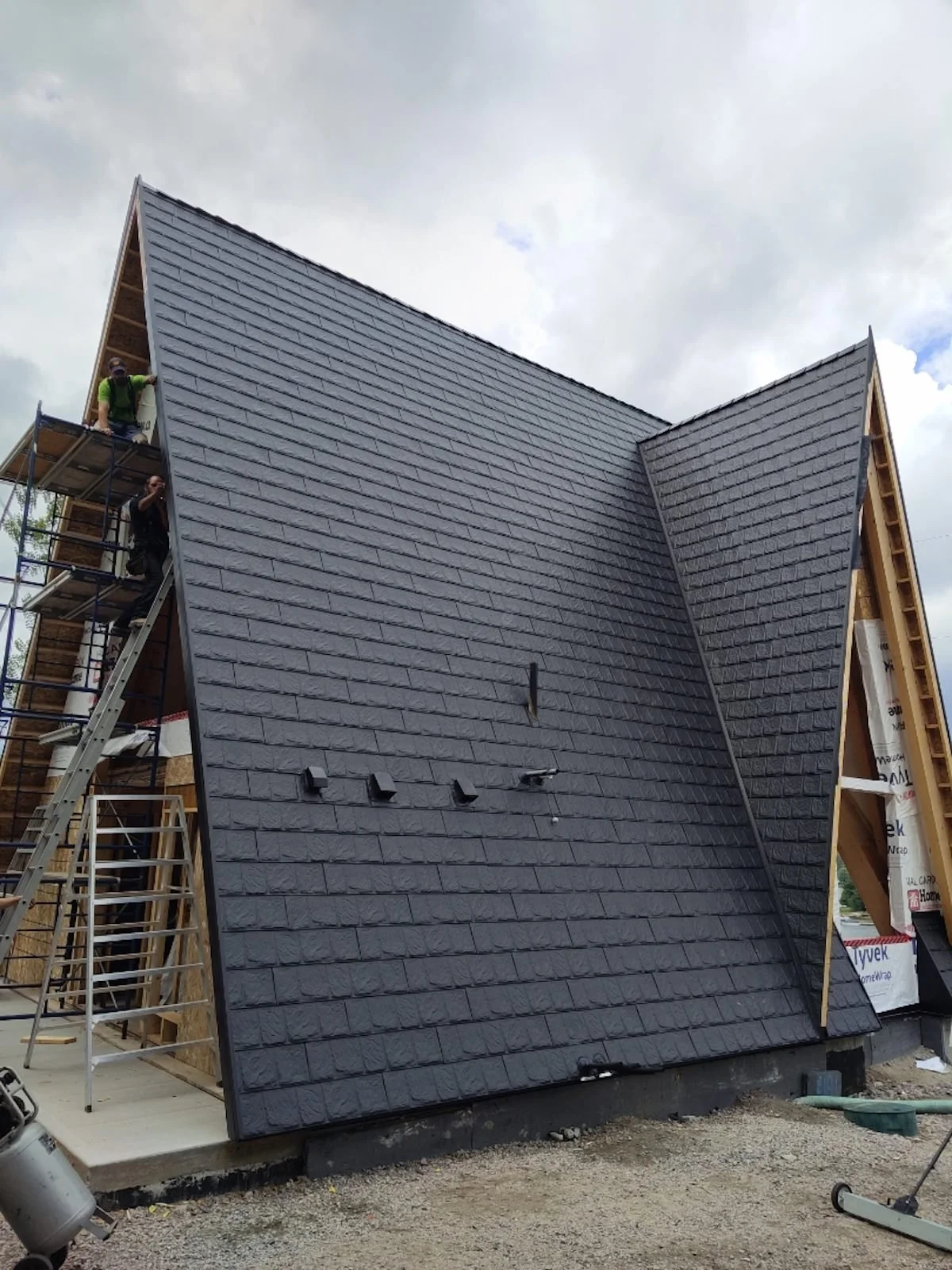 Une maison en construction avec un toit en ardoise noir récemment posé. Des ouvriers travaillent sur une échelle en construction.