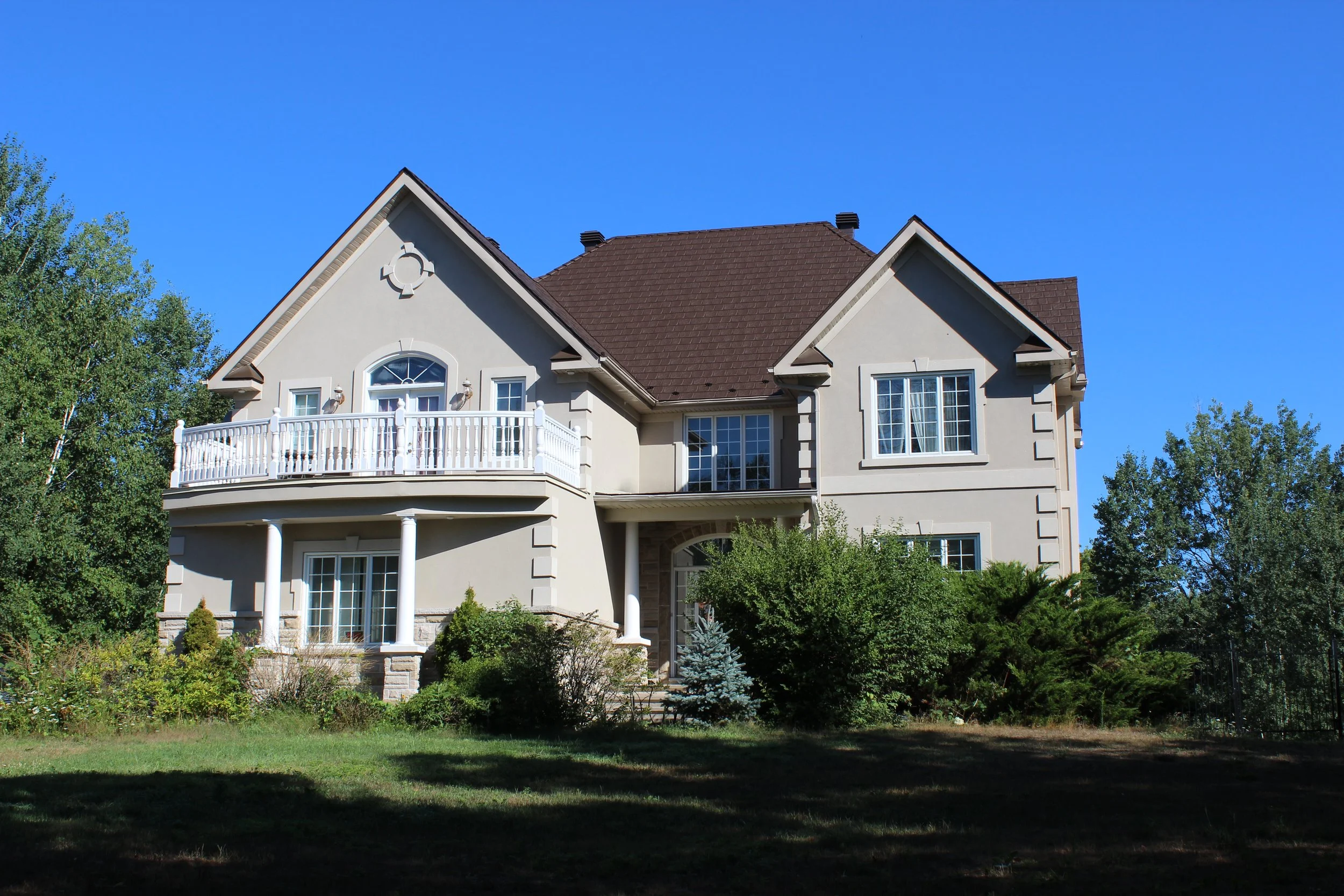 Une grande maison avec un toit en pente, plusieurs fenêtres, une terrasse avec une balustrade blanche et un jardin avec des arbres et des buissons.