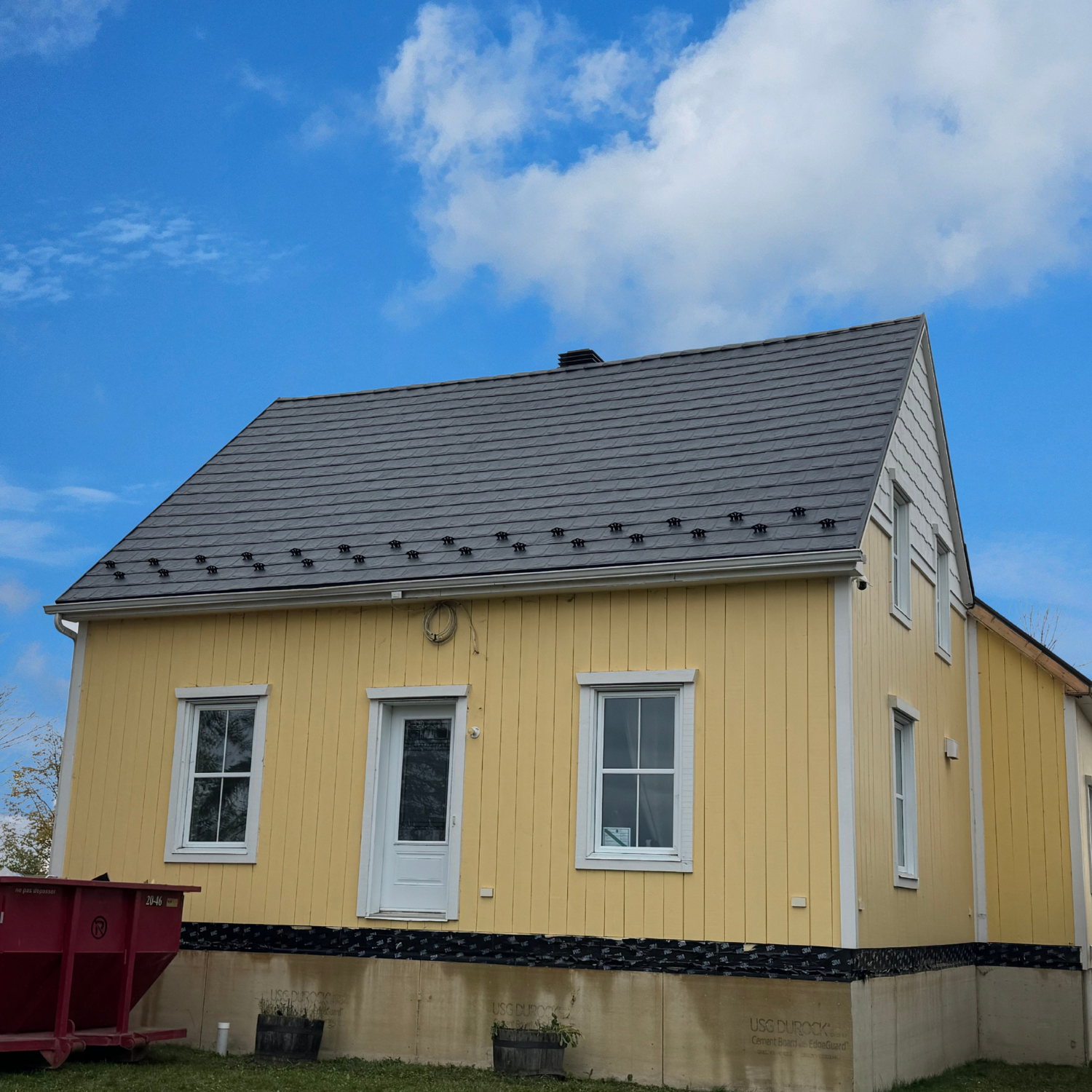 Maison en construction avec façade en bois jaune, fenêtres à double vitrage, toit en tuiles grises, ciel bleu avec quelques nuages.