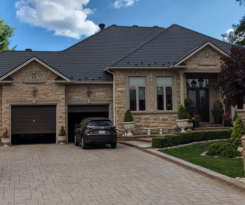 Une maison en pierre avec deux garages, une voiture noire stationnée devant, des fenêtres à volets, un jardin bien entretenu avec des arbustes et des pots de fleurs, sous un ciel bleu avec quelques nuages.