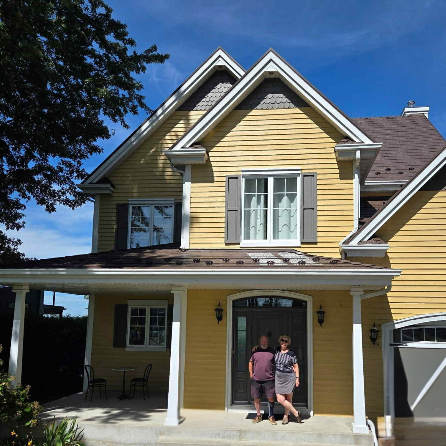 Une maison à deux étages de couleur jaune avec un porche devant l'entrée principale où deux personnes sont debout, en arrière-plan un ciel bleu clair avec quelques nuages, et un arbre à gauche.