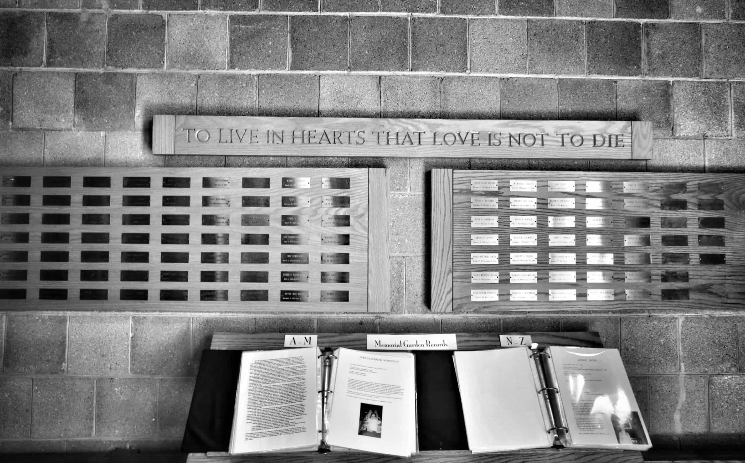 A memorial display with a wooden plaque engraved with the quote, 'To live in hearts that love is not to die,' and two open binders containing records labeled 'A-M' and 'N-Z,' on a brick floor.