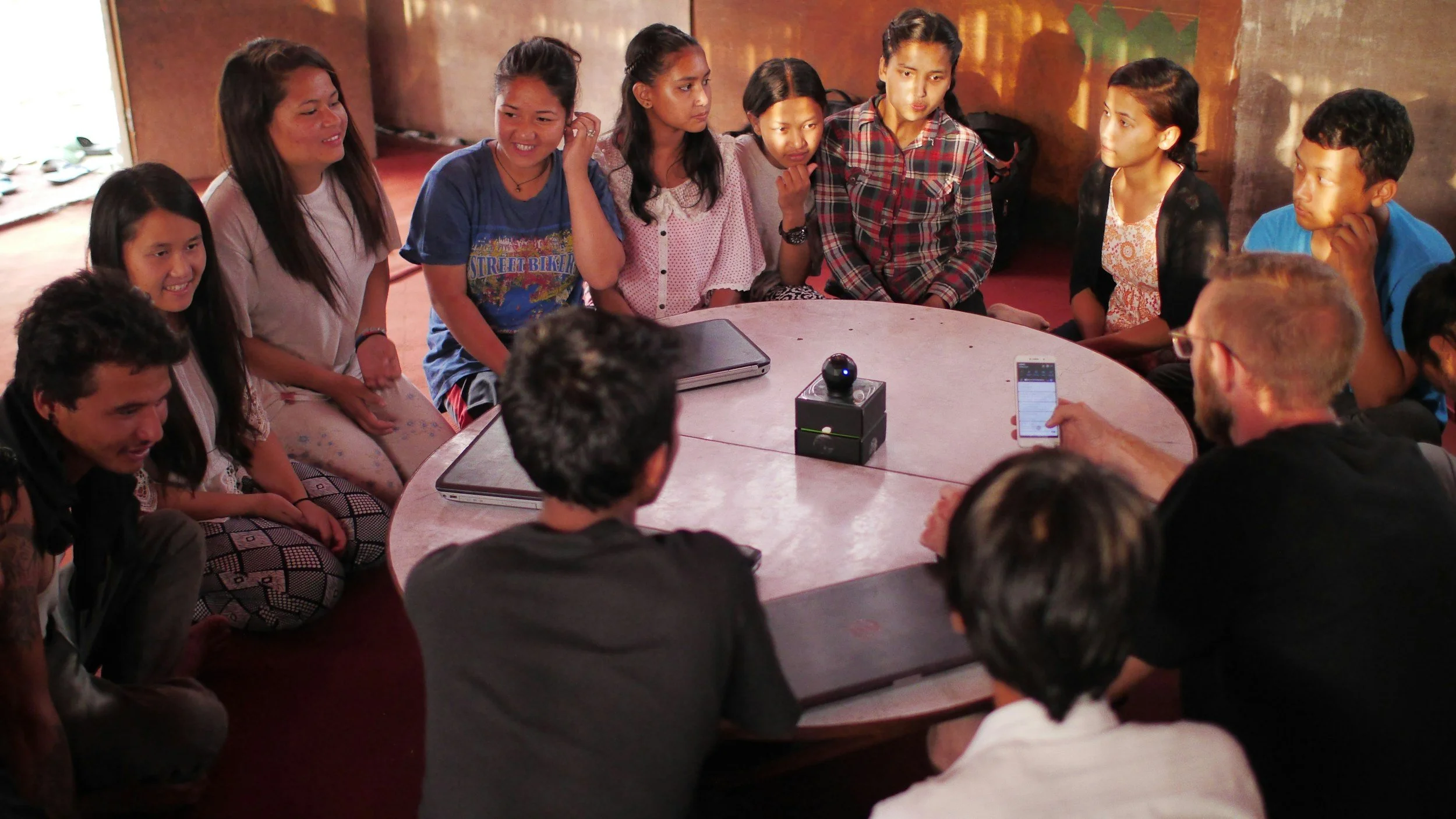 Group of children and young adults sitting and standing around a large round table, engaging in a discussion or workshop, with electronic devices and a small robotic device on the table.