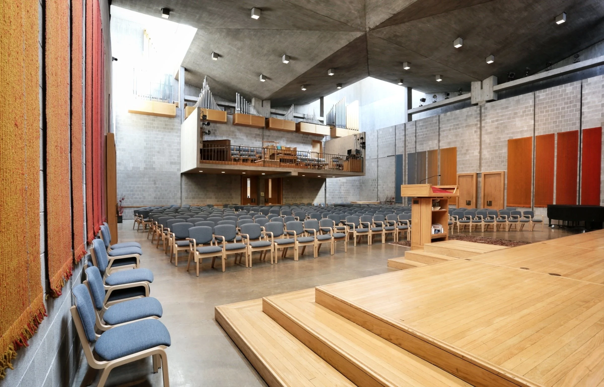 Interior of a modern auditorium or assembly hall with rows of blue chairs, a wooden stage, and a balcony area with musical instruments and lighting. The walls are made of concrete and brick, and the ceiling has embedded lighting.
