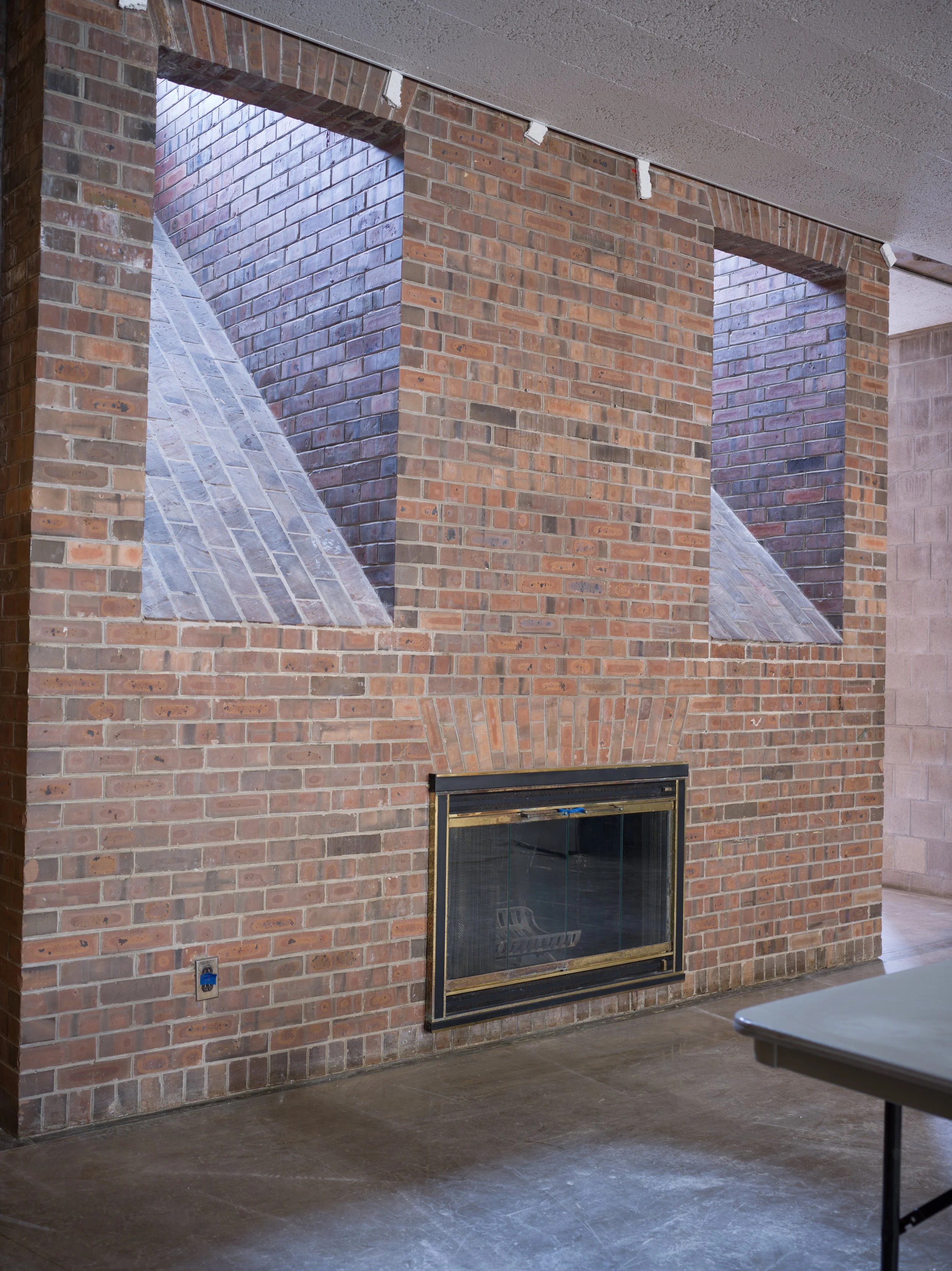 Brick fireplace with a black metal frame, located in a room with concrete floor and a brick wall with three rectangular openings above it.