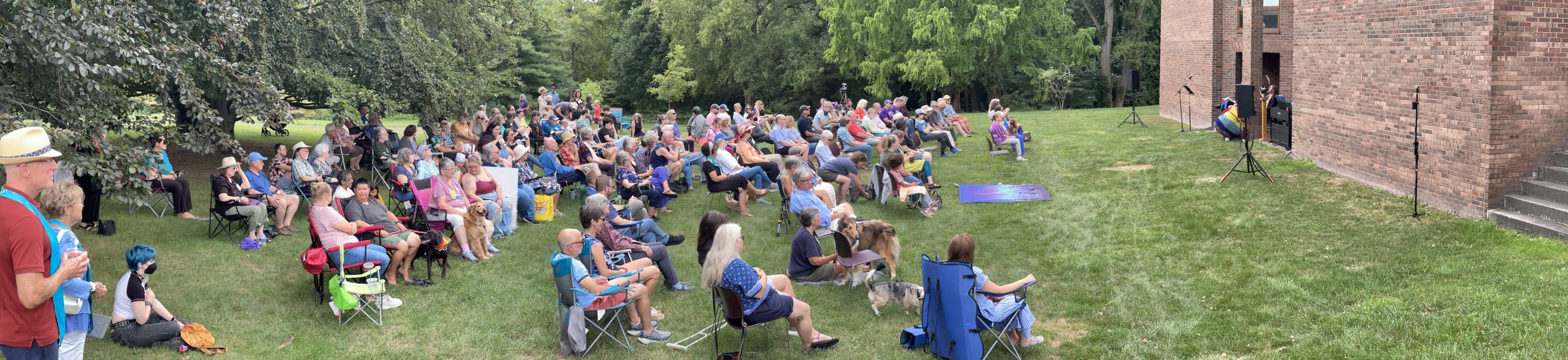 An outdoor event with a crowd of people seated on chairs and blankets on a grassy area, facing a brick building with microphones and speakers set up for a performance or presentation.