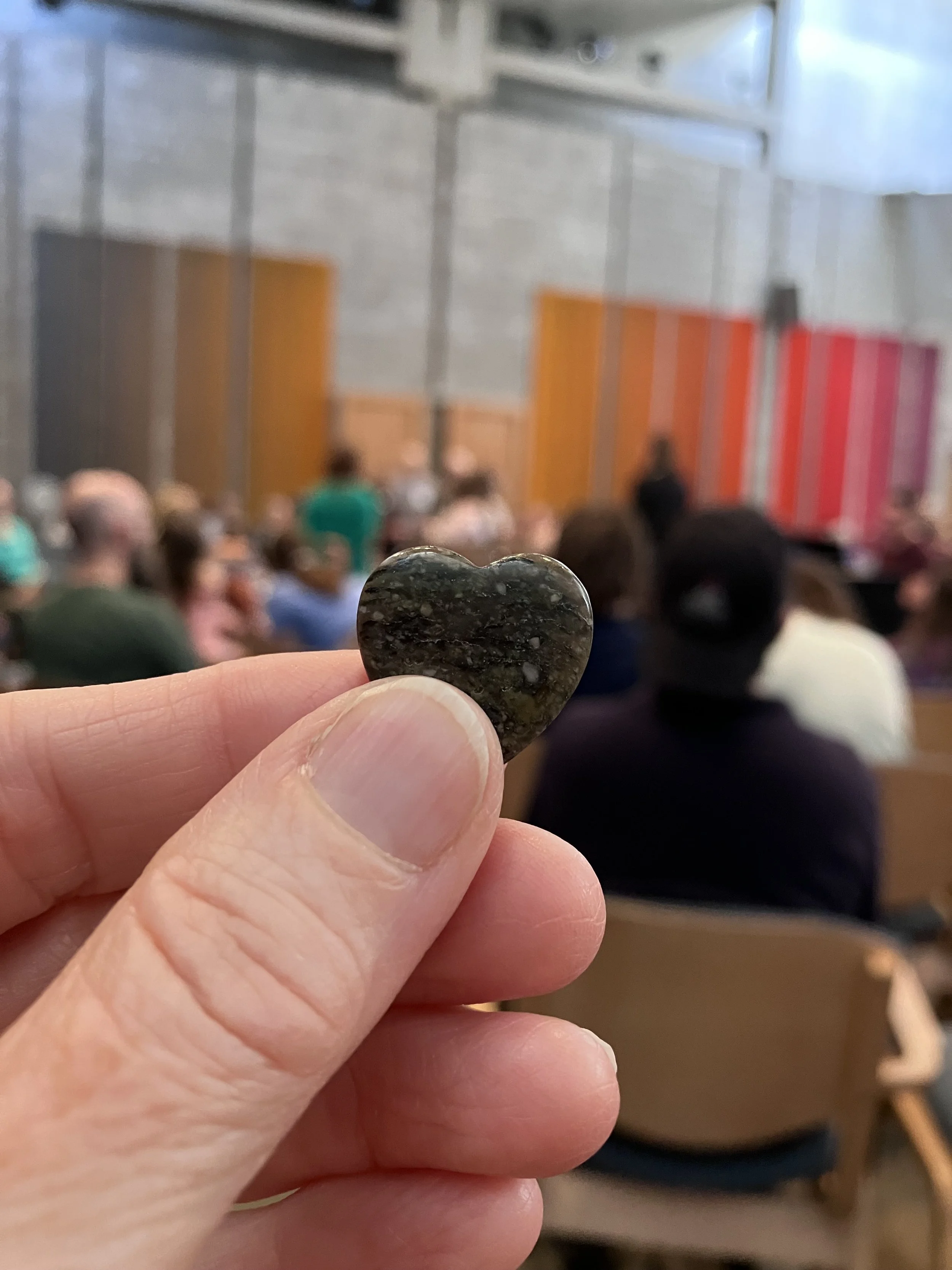 A close-up of a person's hand holding a small, dark, heart-shaped stone. In the background, an audience is seated in a room with colorful wall panels, and a person is speaking at the front.