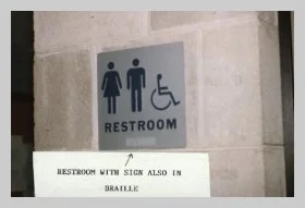 a photo of a sign with an ADA female, ADA male and ADA wheelchair user symbol and the words RESTROOM in all-capital letters as well as in braille. a caption reads "restroom with sign also in braille"