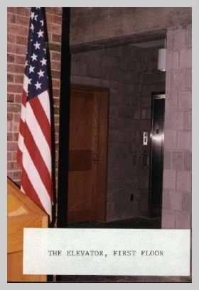 a photo taken in the lobby showing the elevator and American Flag. a caption reads "the elevator, first floor"