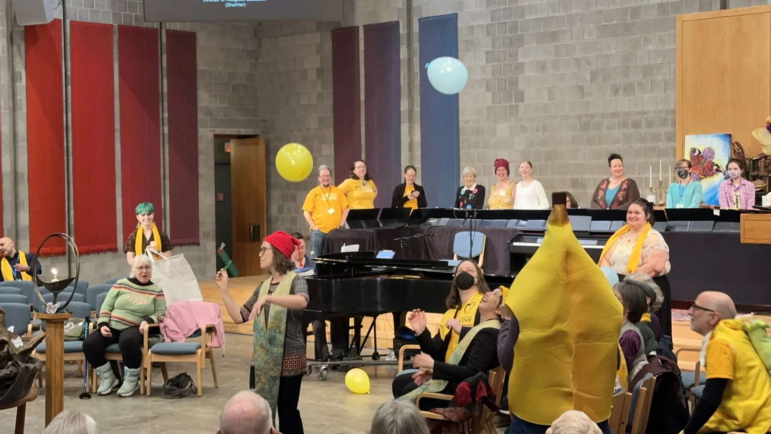 A diverse group of people gathered in a church or community hall decorated for a celebration with yellow balloons and festive attire. Some individuals are standing near a grand piano, while others are seated or dancing. The setting includes a brick wall, colorful banners, and artwork.