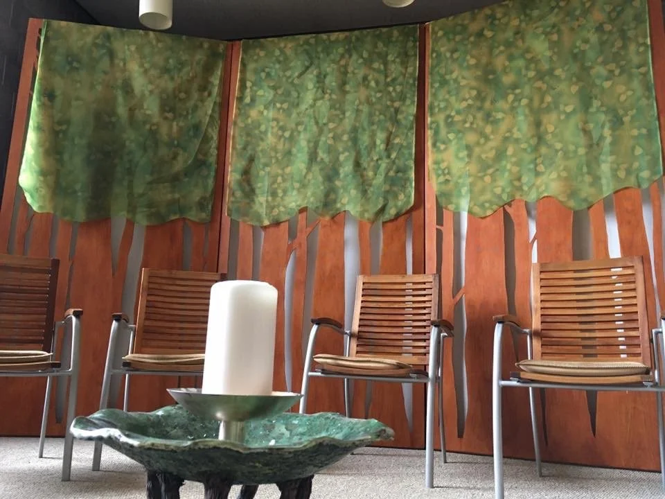 Three wooden chairs with armrests and cushions sit in front of a decorative wooden wall with green fabric curtains hanging above. In front of the chairs, there is a round table with a large greenish stone bowl holding a white candle.
