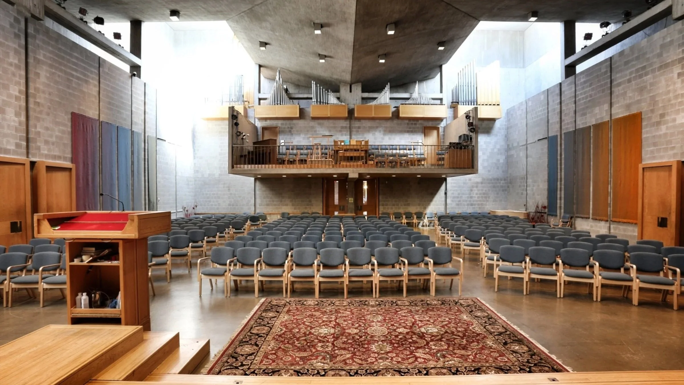 A large, modern church or auditorium interior with rows of gray chairs facing a raised platform with a desk and chairs, a wooden pulpit, and a red patterned rug, warm lighting, and an organ with pipes on the back wall.