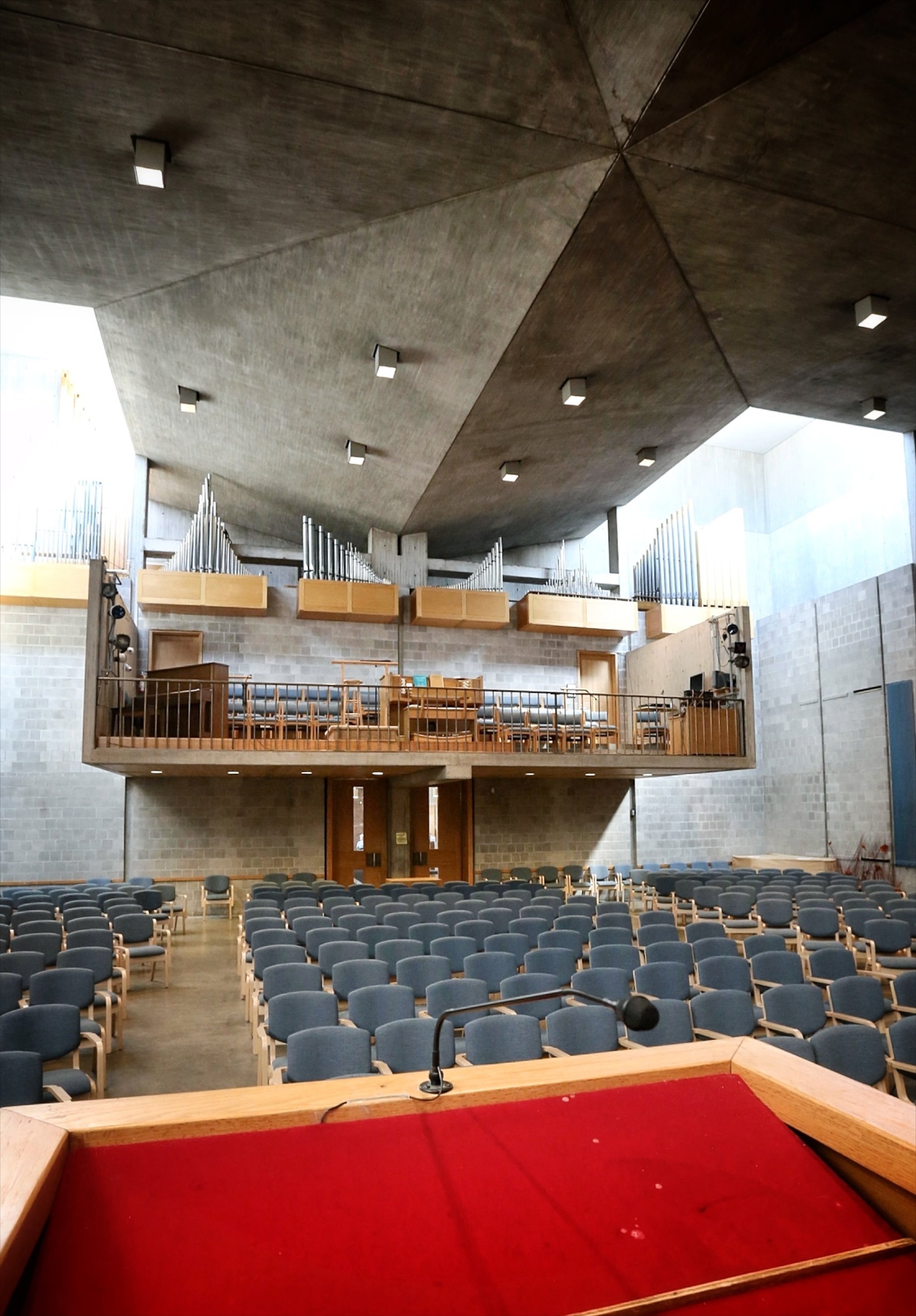 View of an auditorium or conference hall with rows of blue chairs, a stage with red carpet, and a large elevated organ on the back wall. The photo is taken from the podium or stage area.