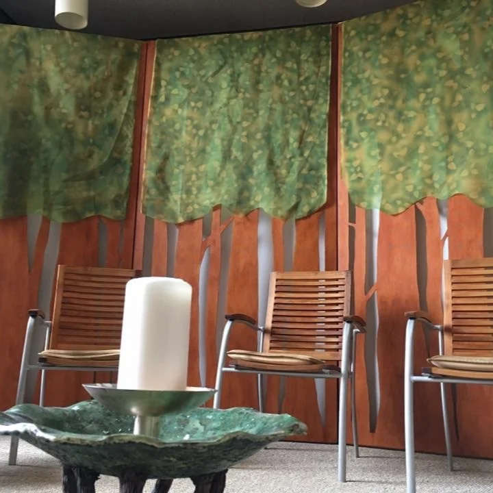 Three wooden chairs with woven seats are placed against a decorative wall with a tree and forest motif and a moss-like green curtain hanging above. A small table with a green, textured bowl-shaped surface holds a large white candle.
