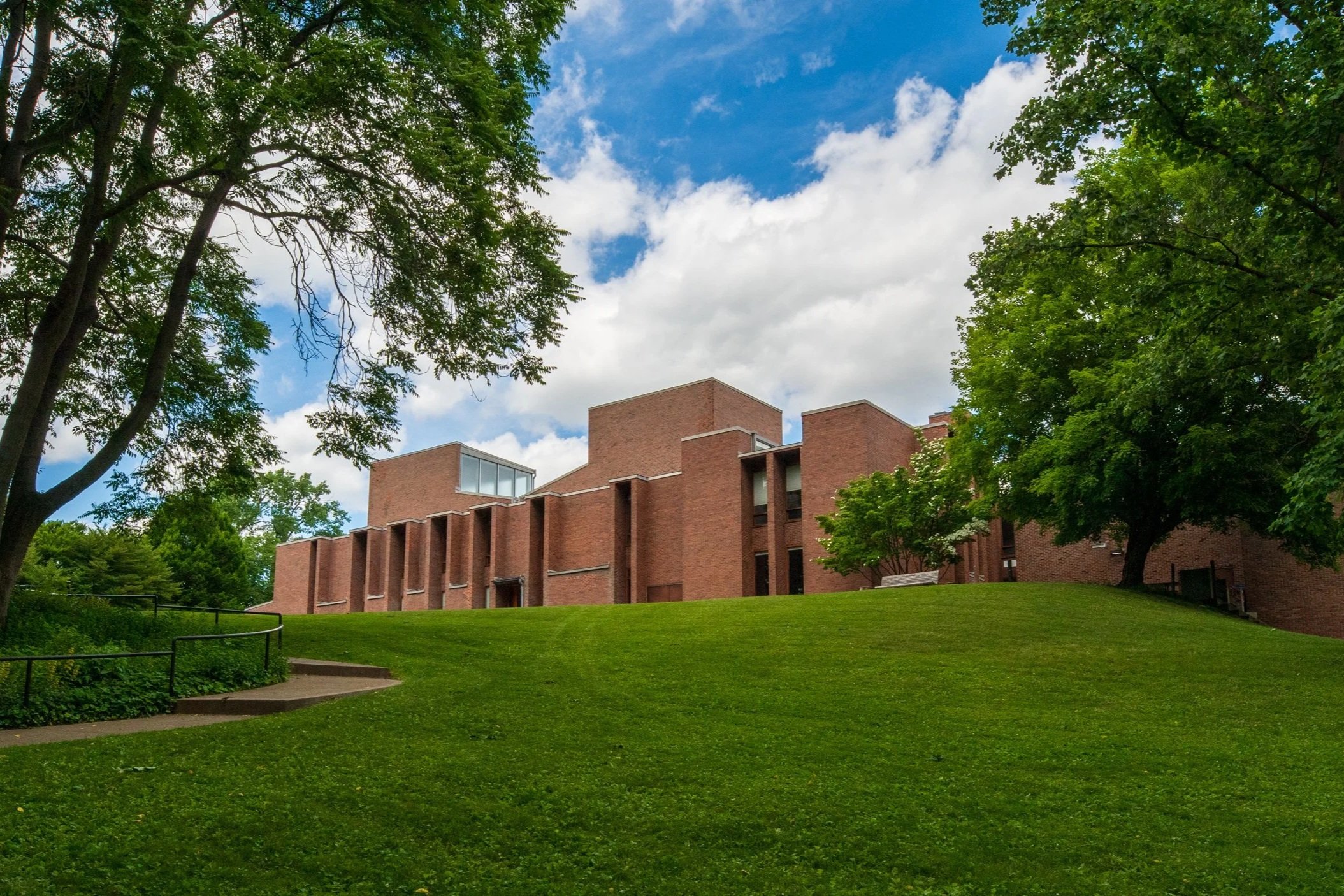 A red brick modern building with angular design, surrounded by green grass and trees with full foliage, under a partly cloudy blue sky.