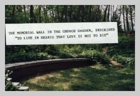a photo of a stone wall in a green garden with tall trees in the background. a caption reads "the memorial wall in the church garden, inscribed 'to live in hearts that love is not to die'"