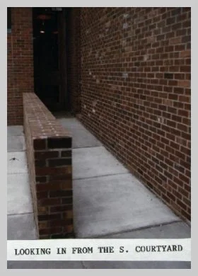 a photo of a walkway with a brick knee-height wall on one side and a brick exterior building wall on the other. a caption reads "looking in from the S. courtyard"