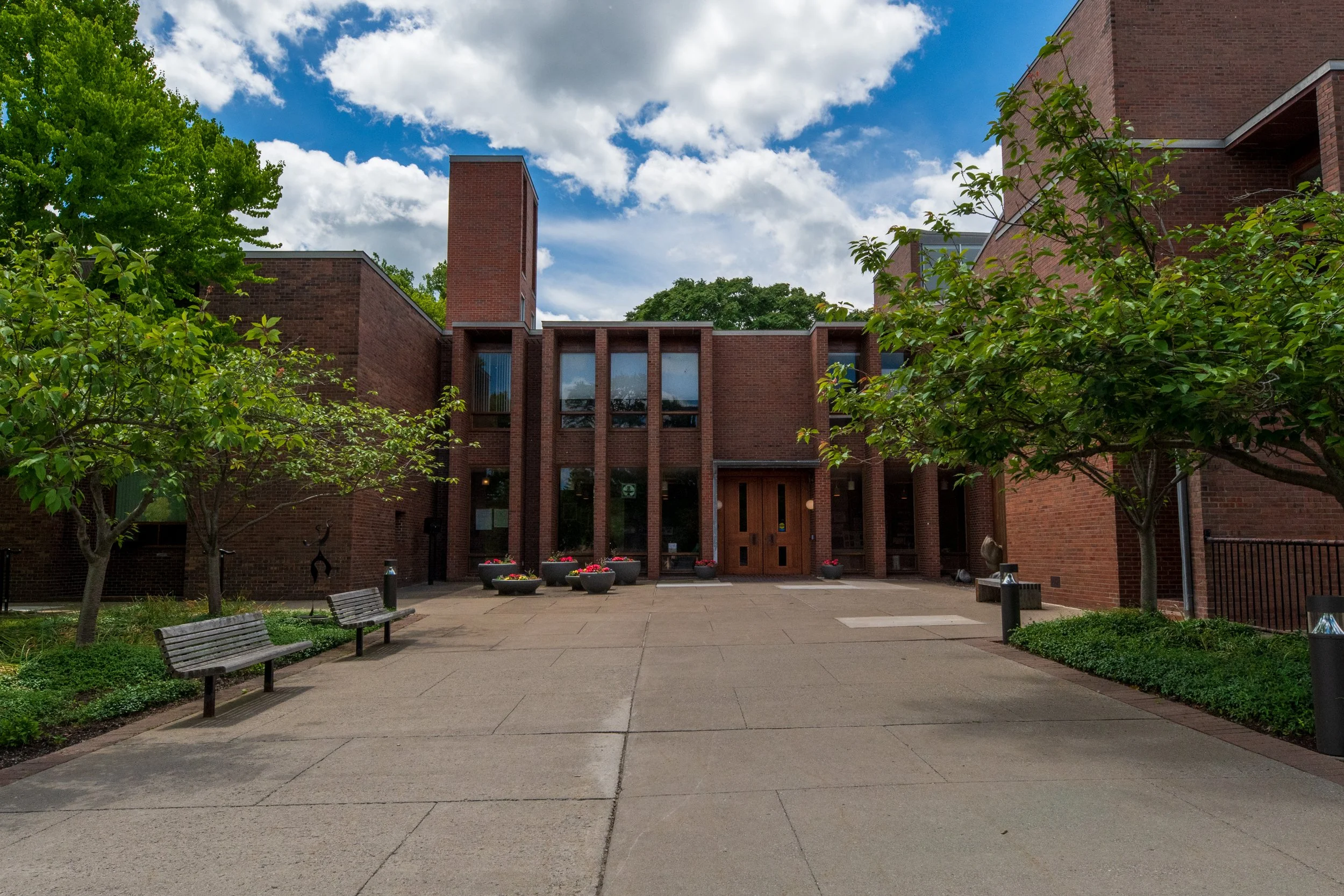 A brick building with trees and blue sky with clouds. In front, there is a paved patio area with benches, planters, and small shrubs.