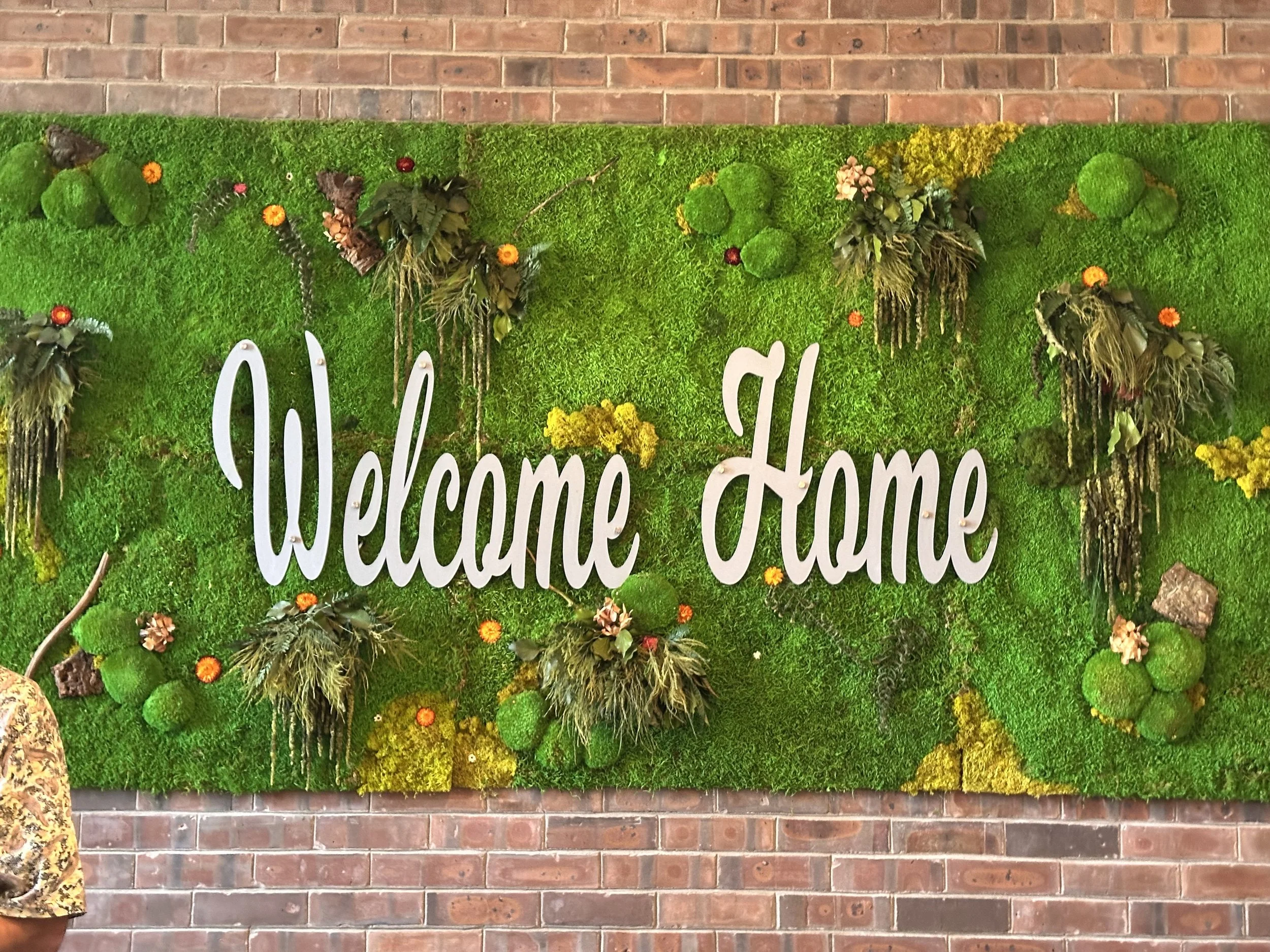 A decorative wall with the words 'Welcome Home' in white cursive letters, surrounded by green moss and small orange flowers, mounted on a brick wall.