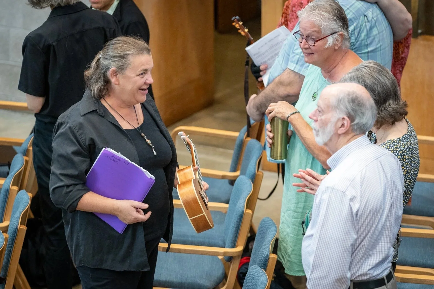 Three people smile and talk in the sanctuary, with one person holding a ukulele.