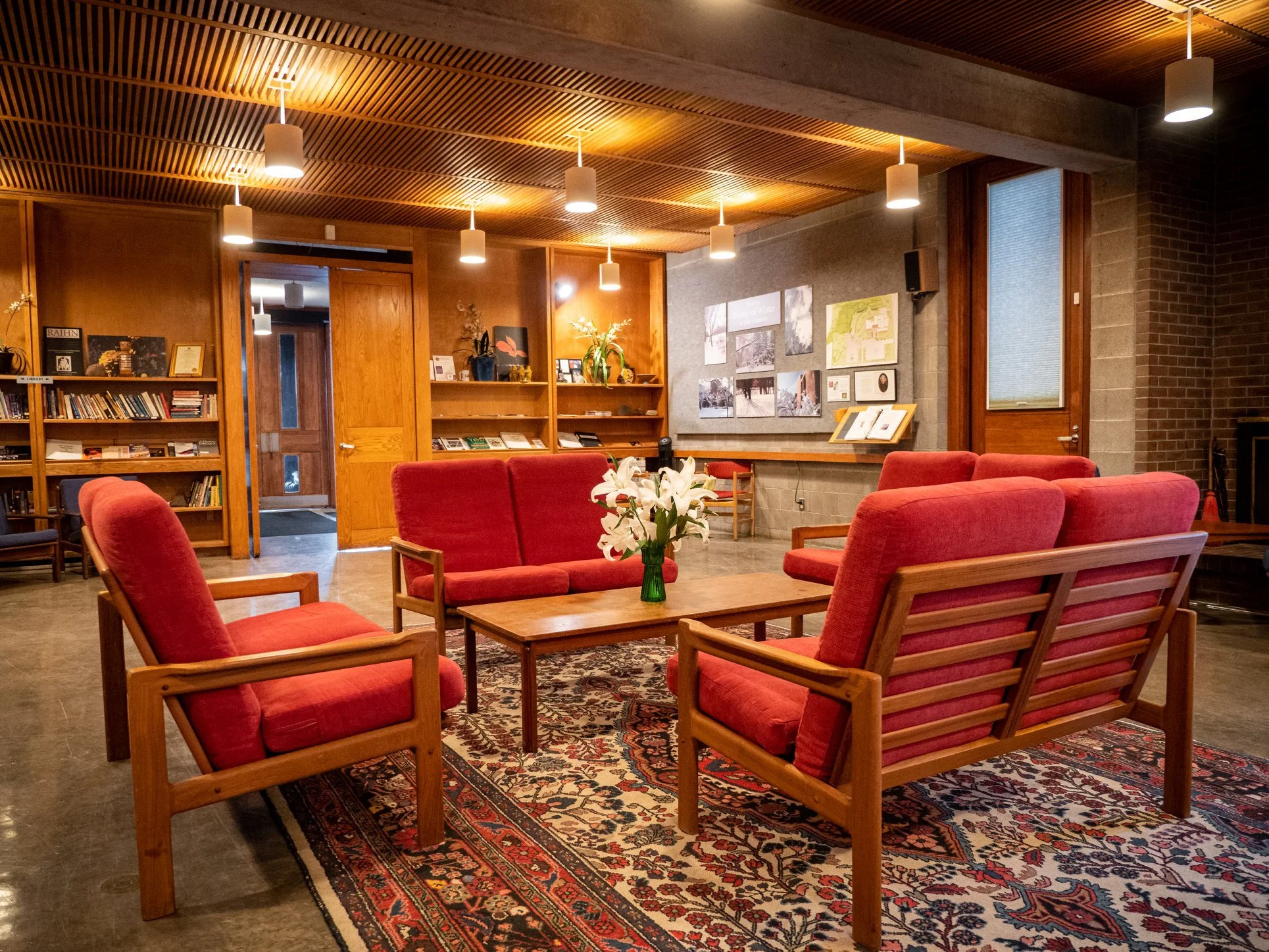 A cozy room with red upholstered armchairs arranged around wooden coffee tables on a patterned rug. The room has wooden ceiling panels with pendant lights, bookshelves, and artwork on the walls.