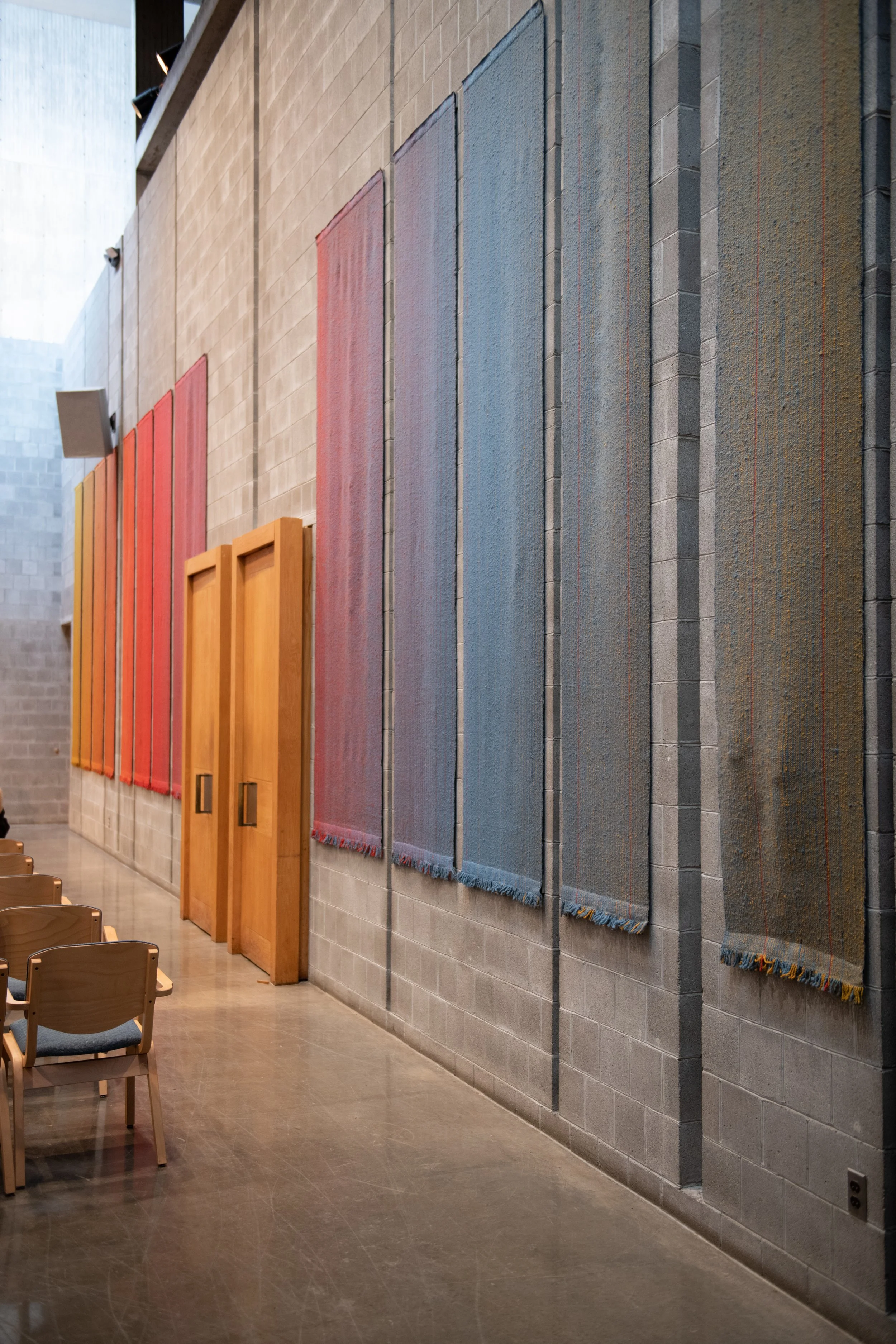 Interior of a room with a light-colored brick wall decorated with several hanging woven textiles in red, blue, and beige, and two wooden lecterns, with chairs lined up in front.