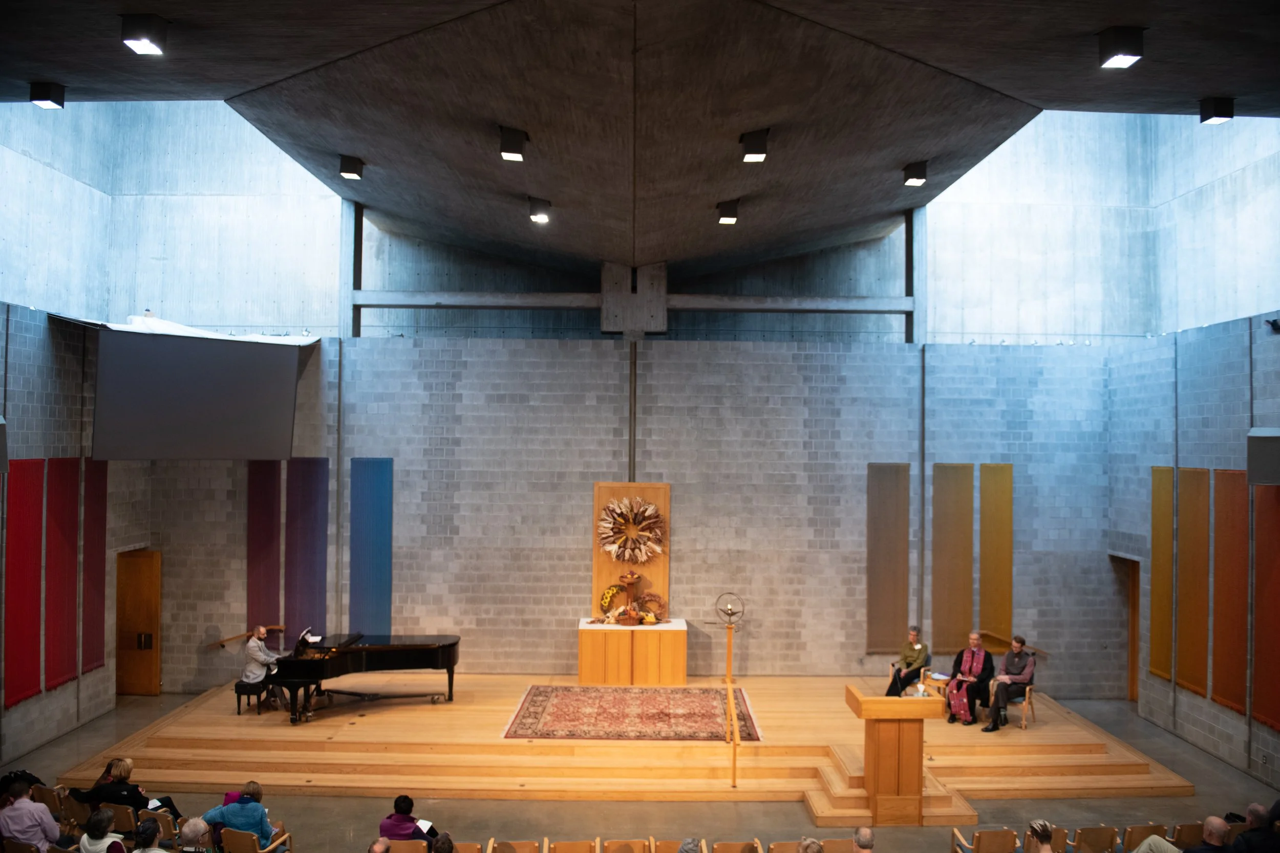 Interior of a church or auditorium with a wooden stage, an altar with a religious wreath, a pianist playing a piano, and a few people seated, some wearing purple and pink robes.