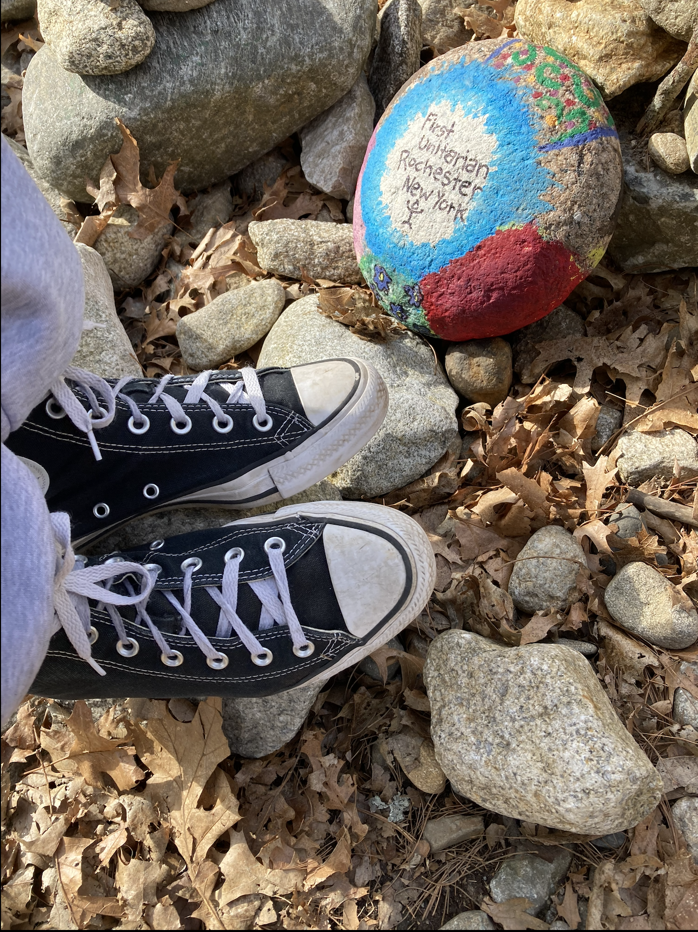 Person wearing black and white sneakers standing on rocky ground with dried leaves, next to a painted rock that says "High Unitarian Richenster, New York."