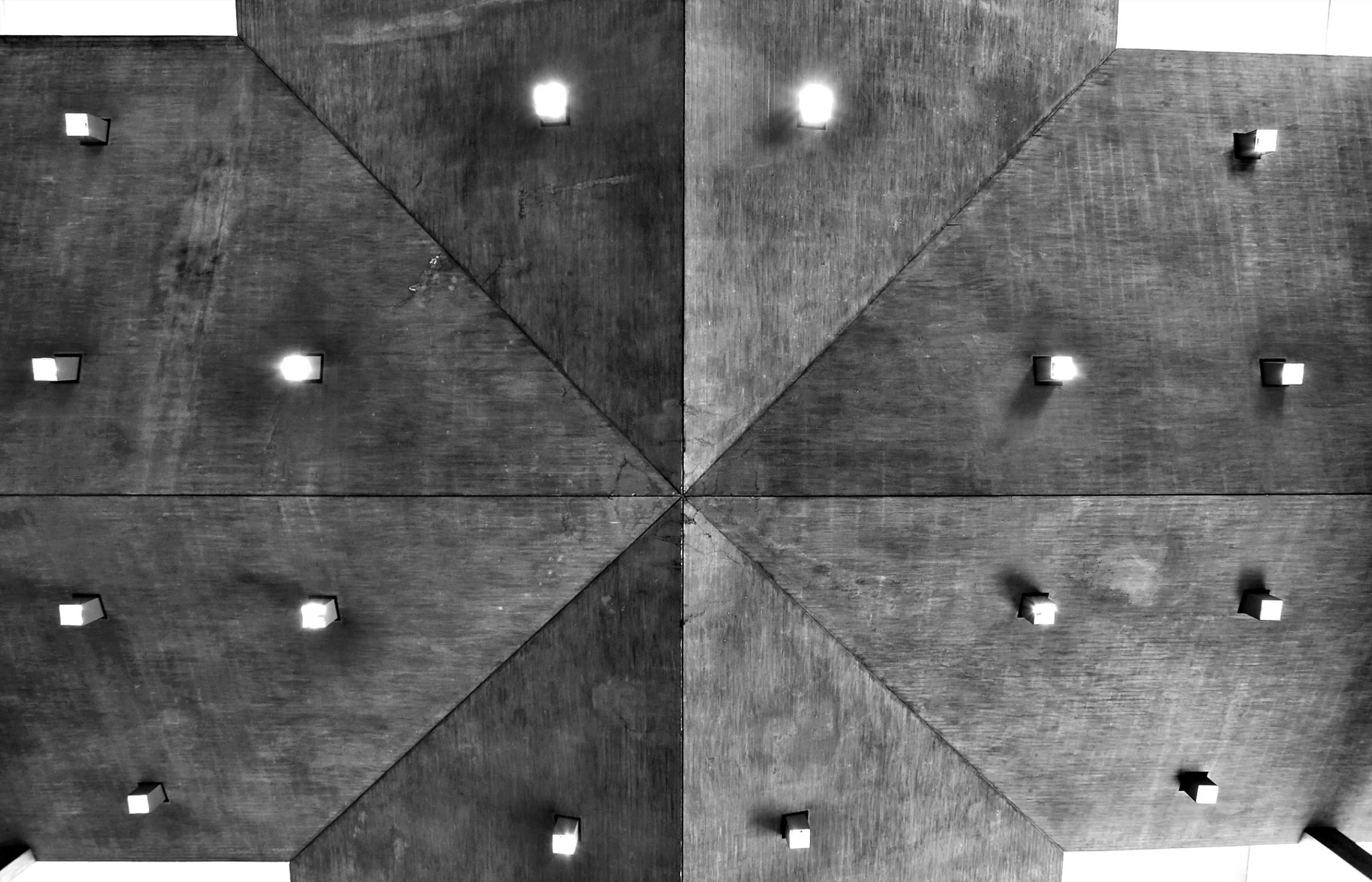 A ceiling with a geometric pattern of wooden beams forming a triangular shape, with small square recessed lights evenly spaced along the beams.