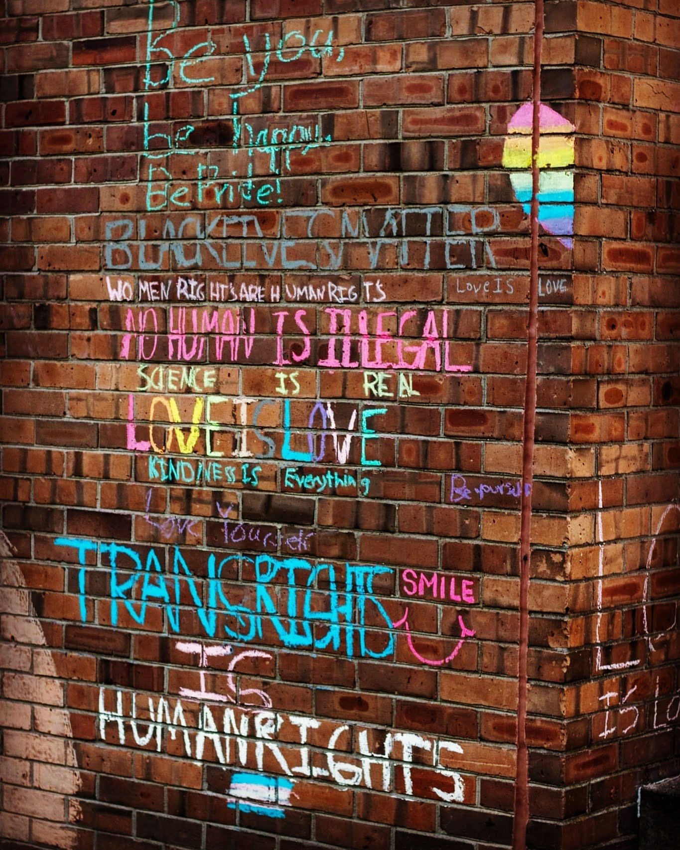 A brick wall covered with colorful chalk messages including “Love is love,” “Science is real,” and “Human rights.”