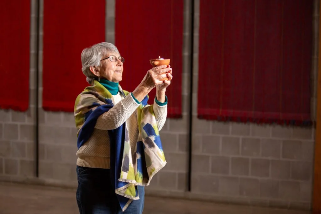 An elderly woman with gray hair and glasses holding a lit candle inside a building with cinderblock walls and colorful wall panels.