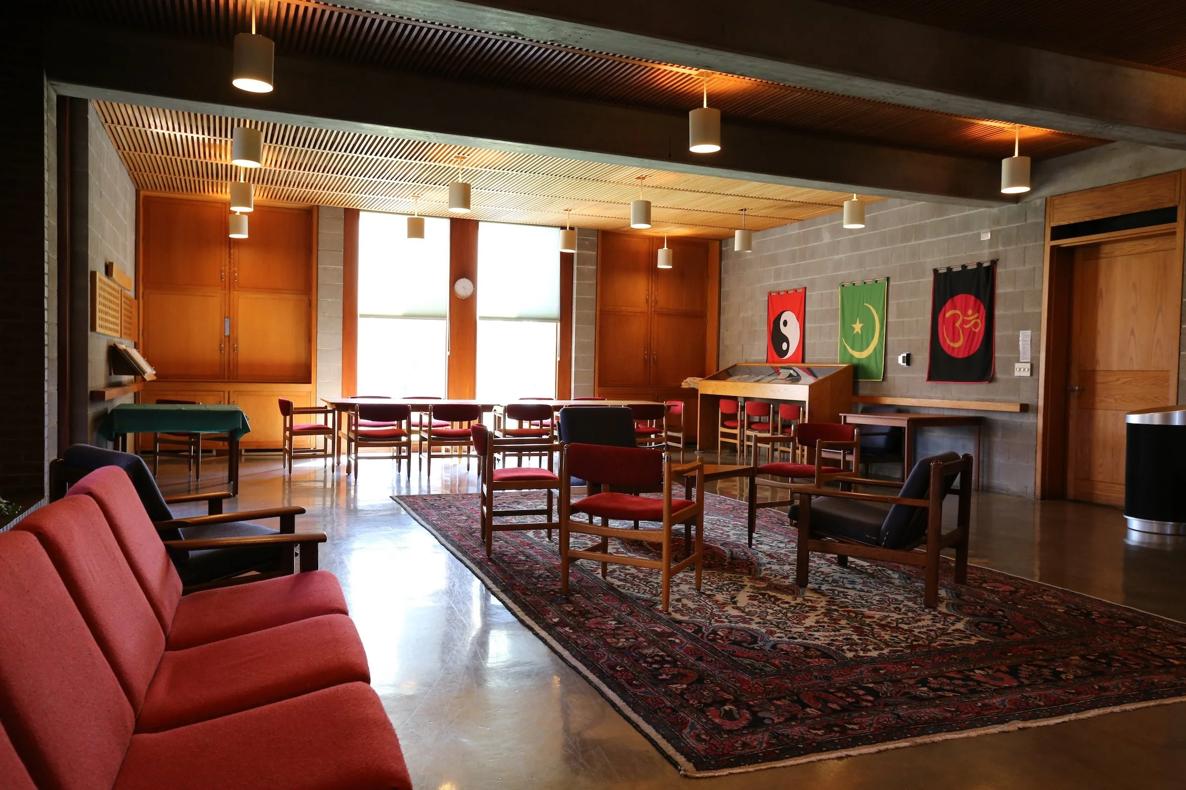 A cozy room with red and black chairs, a patterned area rug, wooden cabinets, and wall hangings with symbols, illuminated by pendant lights and large windows.