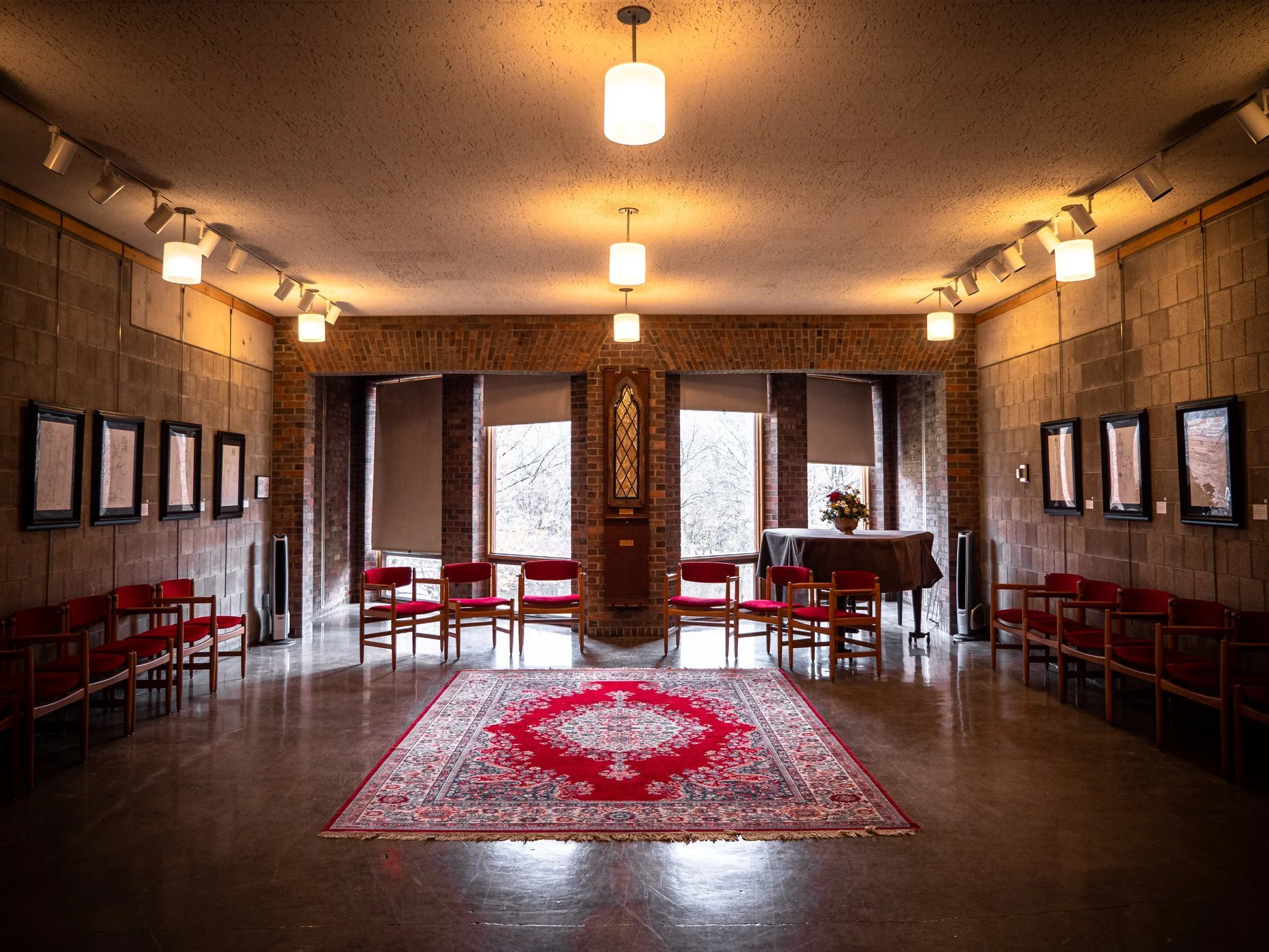 An interior with brick and stone walls, large windows, red chairs arranged in a semi-circle, a floral arrangement on a table, and a red patterned rug on a polished floor, lit by ceiling lights.