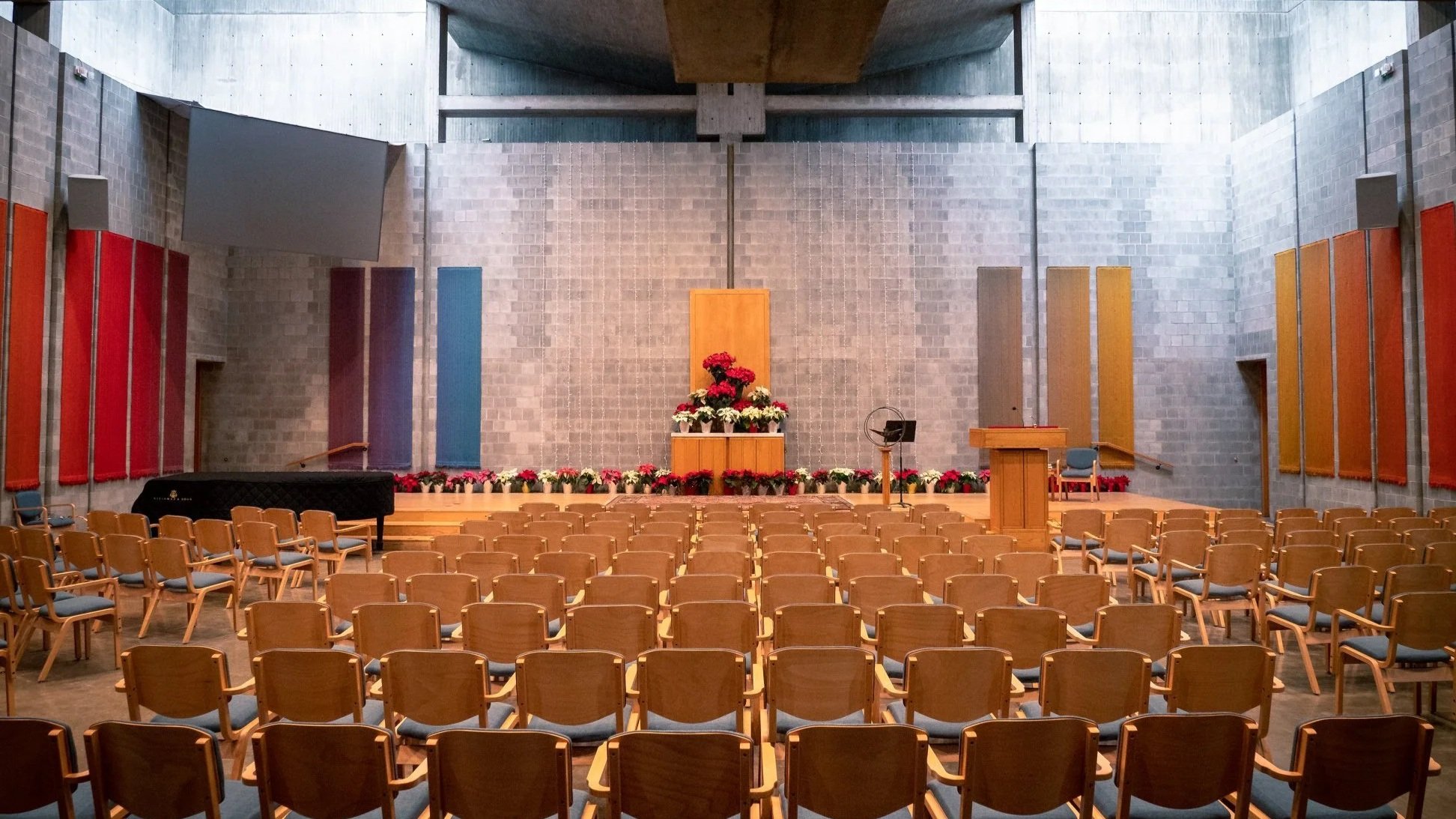 Interior of a church or auditorium with wooden chairs arranged in rows facing a stage. The stage features a floral display at the center, a wooden podium, a chair, a music stand, and a piano. The wall behind the stage is made of gray bricks with colo