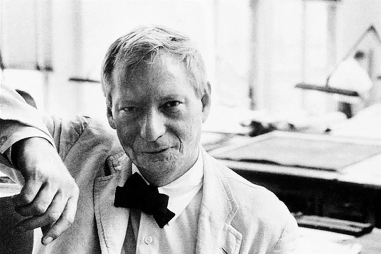 Black and white photo of a man with light hair and a bow tie, leaning on a counter or table in an indoor setting, with office or workspace background.
