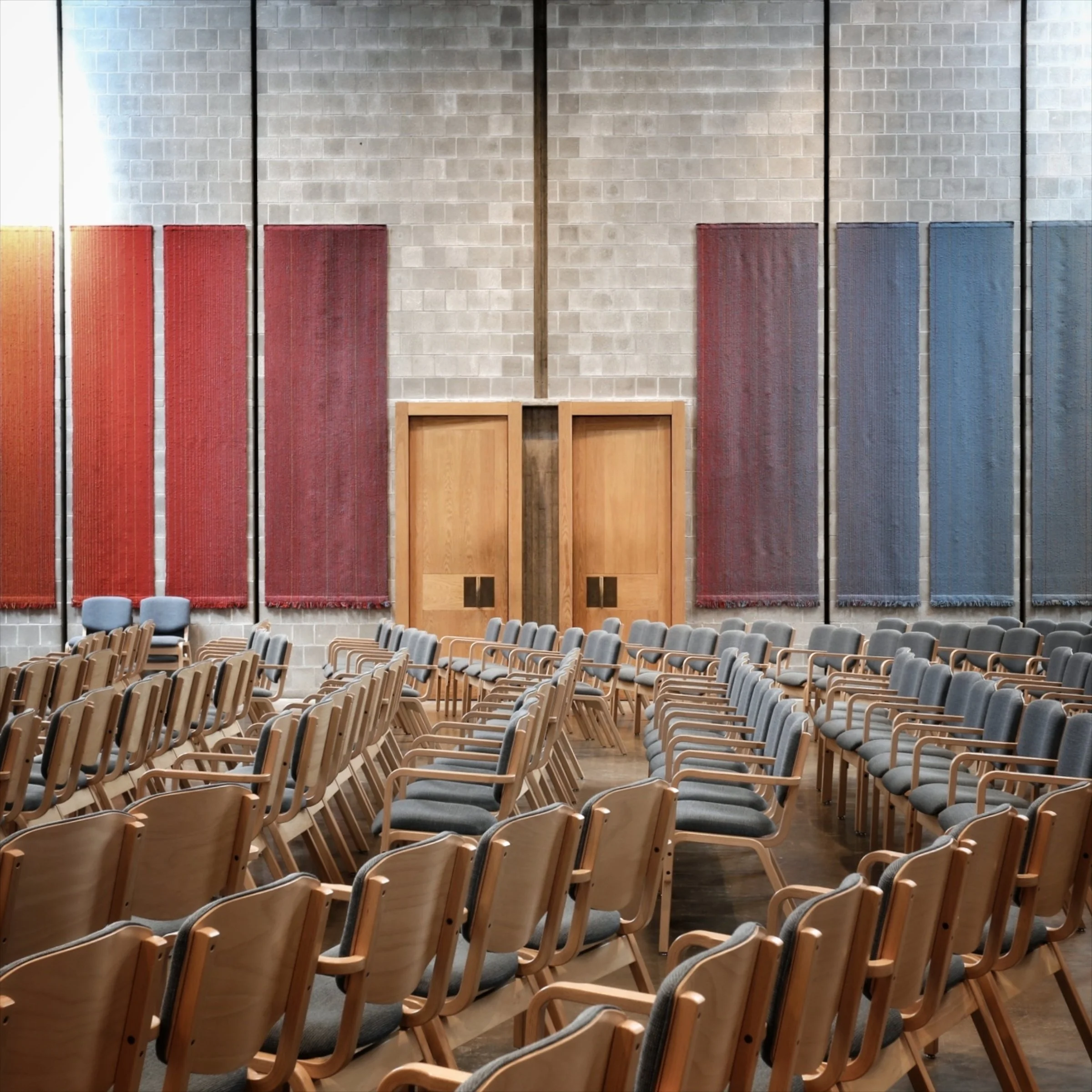 An empty auditorium with rows of wooden chairs with padded seats facing a stage area. The backdrop features a large grey brick wall adorned with four tall, narrow, colorful hanging fabric banners in red and blue, separated by wooden beams.