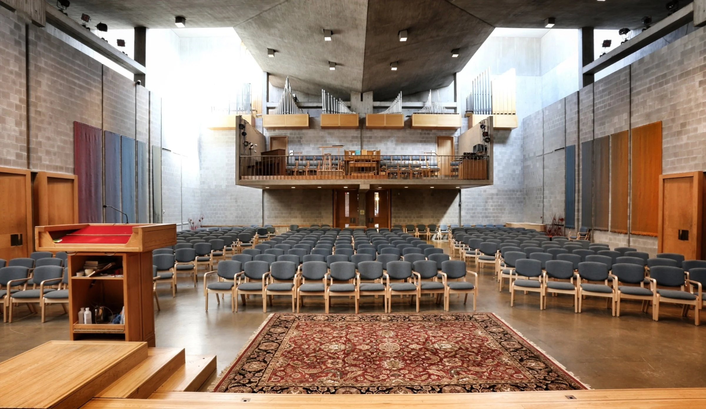 Inside a large modern church or concert hall with many gray chairs arranged in rows facing a wooden pulpit with a red carpet, and a balcony with a pipe organ and musical instruments at the back.