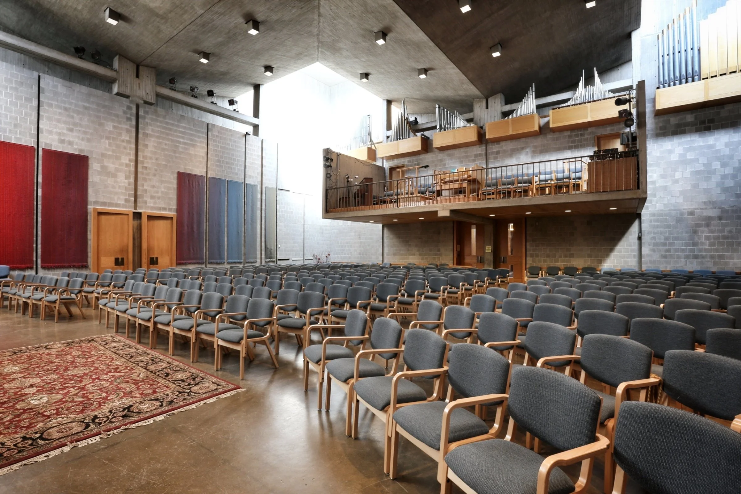 An empty concert hall with rows of chairs, a stage, and a balcony with musical pipes, illuminated by natural light from large windows.
