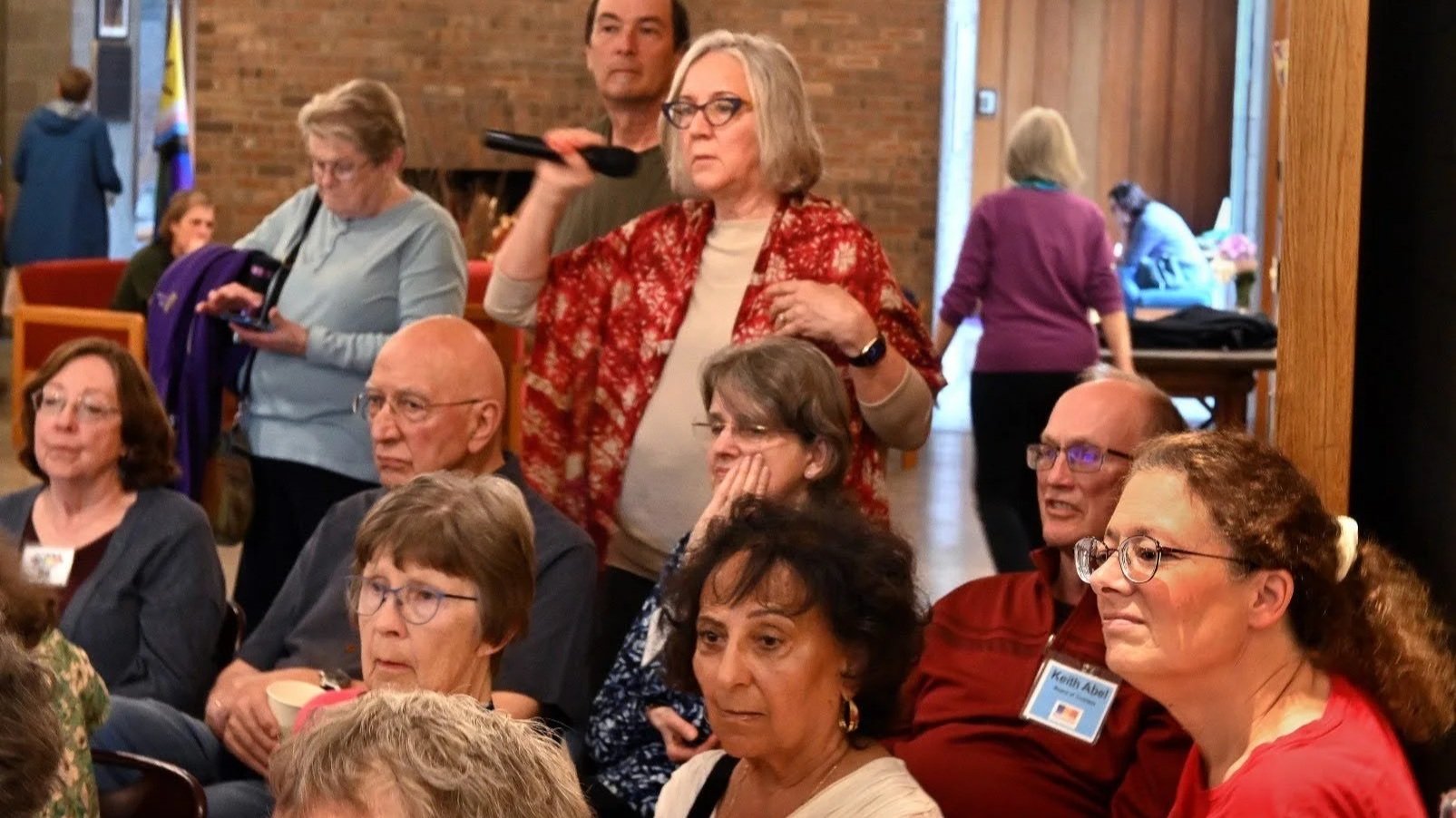 Several older adults seated in a room, some looking attentive, one woman standing holding a microphone, some women walking in background, brick walls, and wooden pillars.