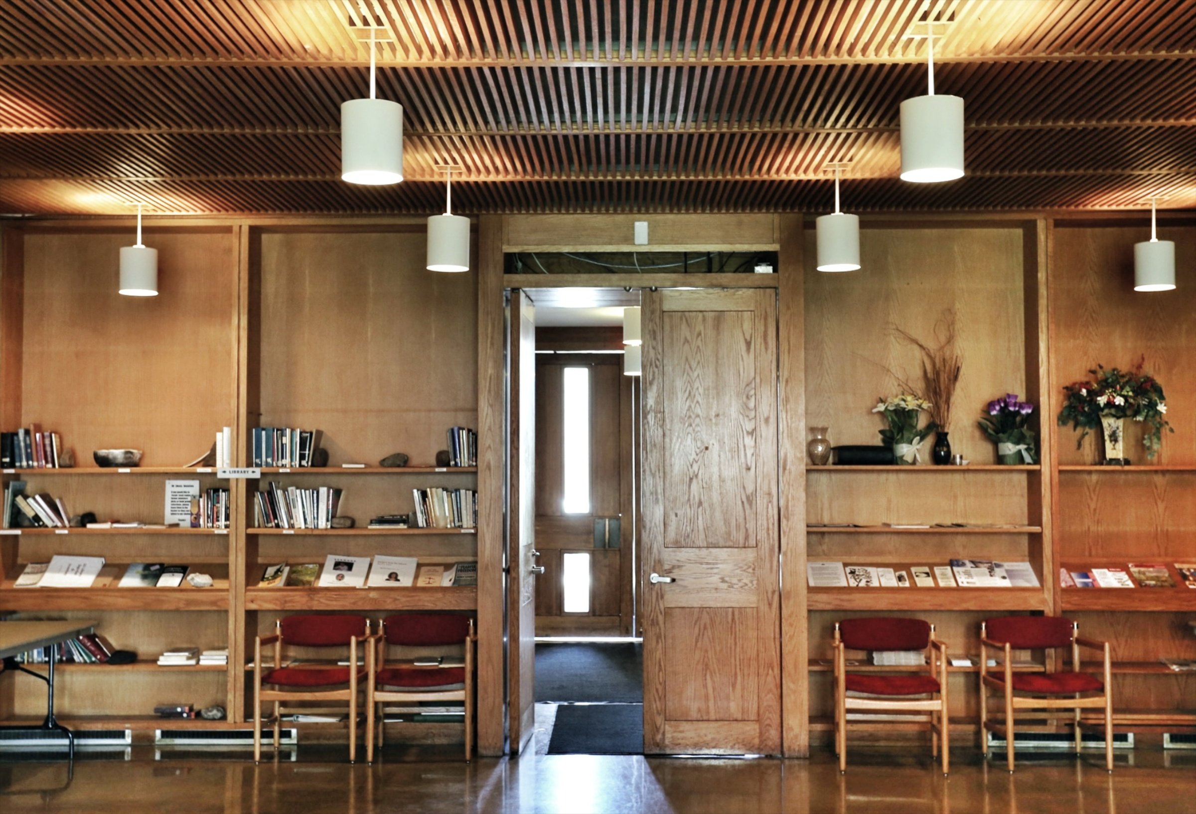 Indoor space with wooden panel walls, bookshelves, and chairs, bright lighting from ceiling lamps, partially open door leading to another room, and decorative plants and flowers.