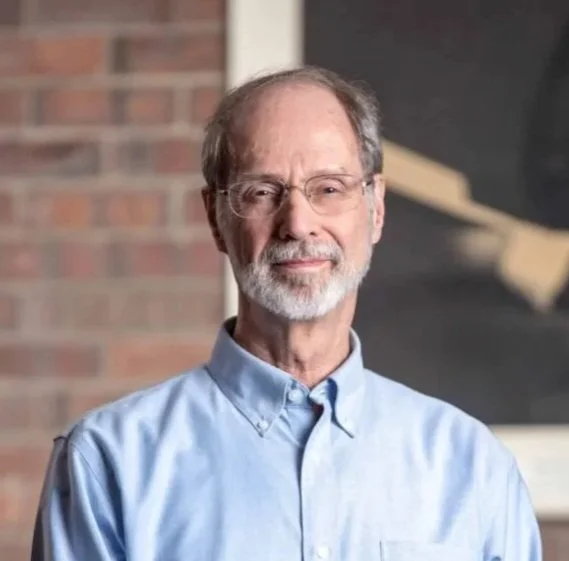 Portrait of an older man with glasses and a short gray beard wearing a light blue button-down shirt, standing in front of a brick wall and artwork.