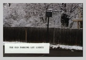 a photo of a snow-covered light post, fence and hedge with a caption that reads "the old parking lot lights"