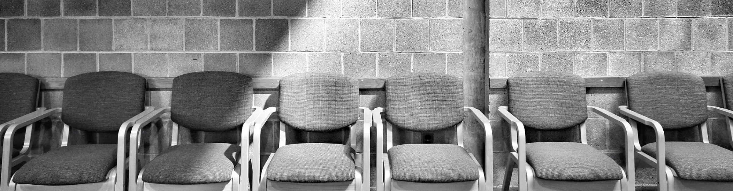 Eight empty chairs with gray upholstered seats and backs, arranged in a row against a brick wall in a waiting area.