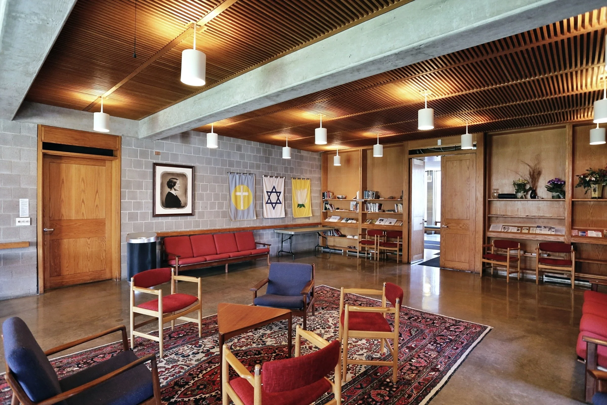 Interior of a lounge or waiting area with wooden ceiling, pendant lights, framed portrait, flags, bookshelf, wooden furniture, and patterned rug.