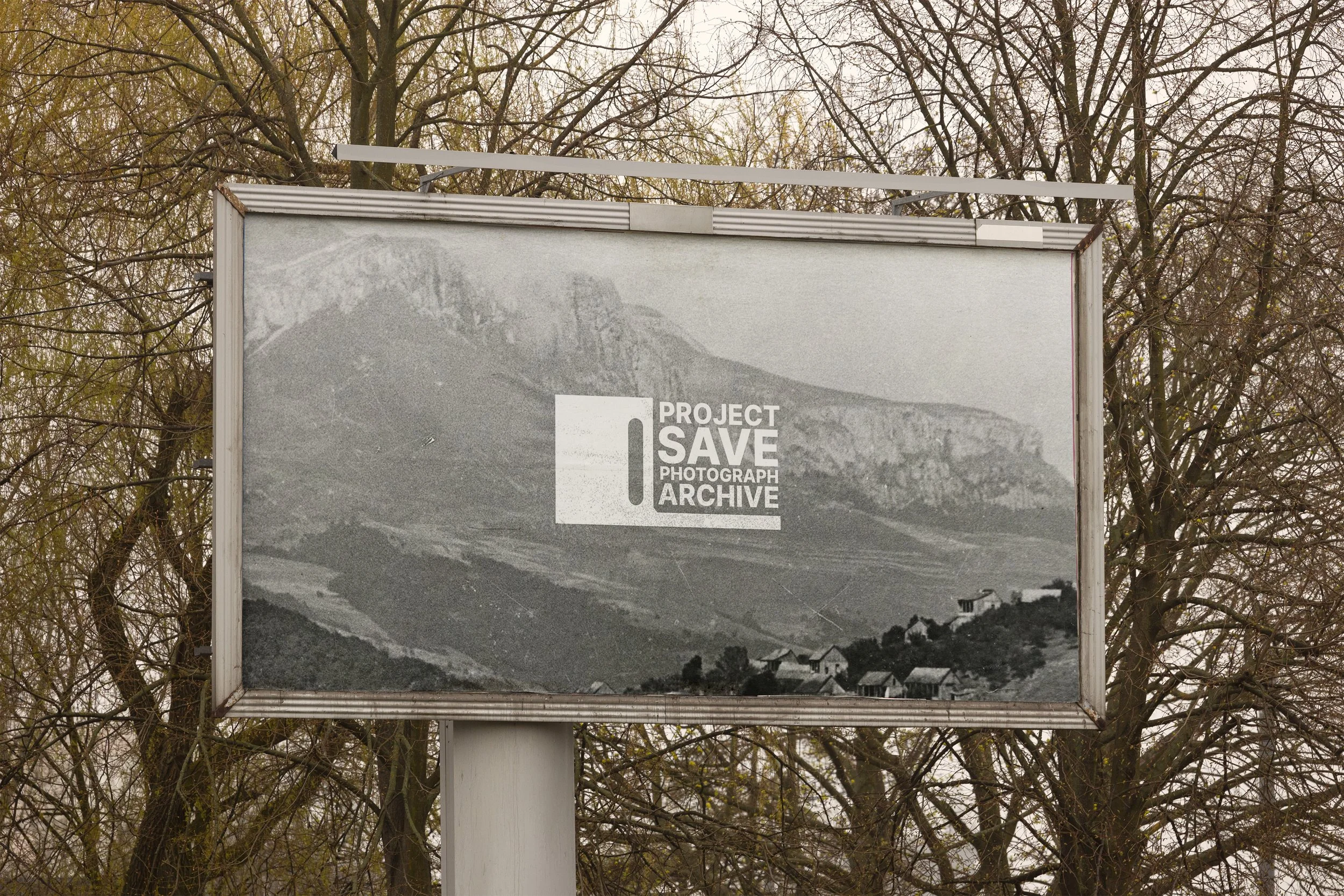 A billboard with a black and white photograph of a mountainous landscape, featuring a town at the base of the mountain and the text 'PROJECT SAVE PHOTOGRAPH ARCHIVE' in the lower right corner, set against a background of leafless trees.